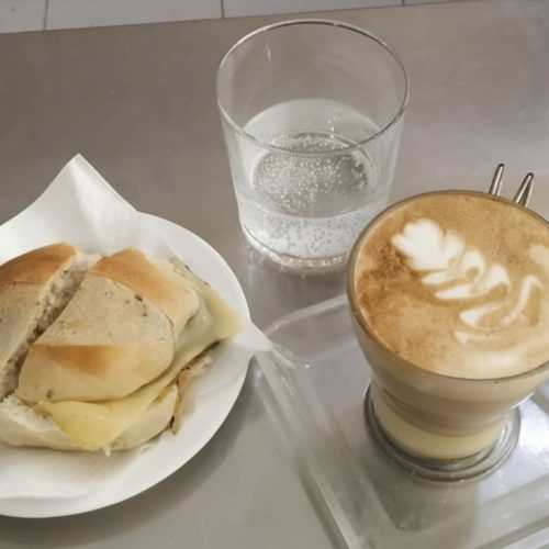Un bocadillo de carne con queso en un plato, junto a un vaso de agua con gas y una taza de café con leche con arte latte en forma de hoja, todo dispuesto sobre una mesa de acero inoxidable.