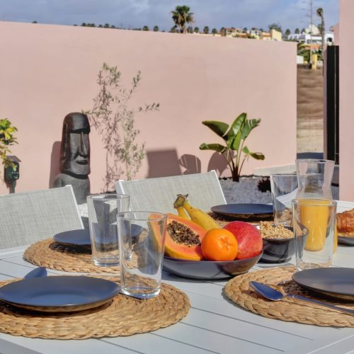 Mesa exterior servida con fruta, zumo y vajilla; pared rosa y figura decorativa