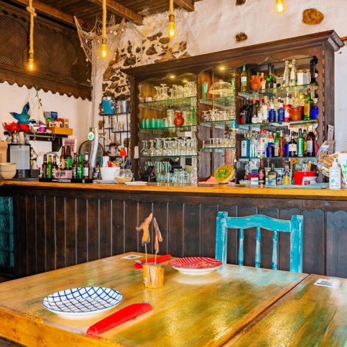 Comedor con mesas de madera, sillas turquesa y barra con botellero iluminado en A Mi Manera.