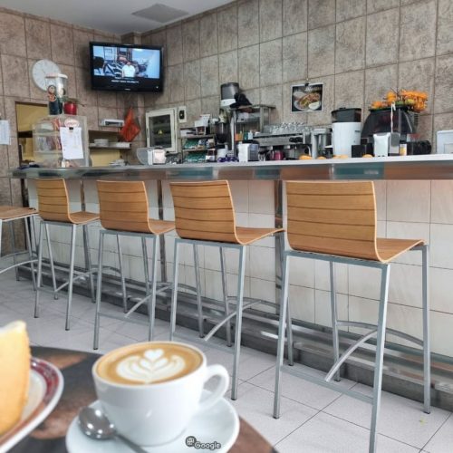 Interior de la "Cafetería del 15" en Puerto del Rosario con una barra larga y seis taburetes de madera, una televisión montada en la pared y una taza de café con arte latte en primer plano.