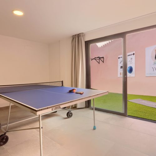 Mesa de ping-pong junto a ventanal con salida a patio de césped y carteles de yoga