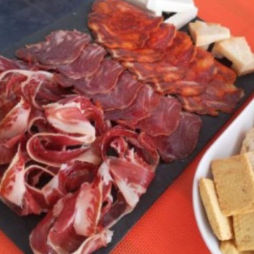 Tabla de embutidos ibéricos variados, incluyendo jamón, lomo y chorizo, servida en una pizarra junto a trozos de queso y picos de pan.